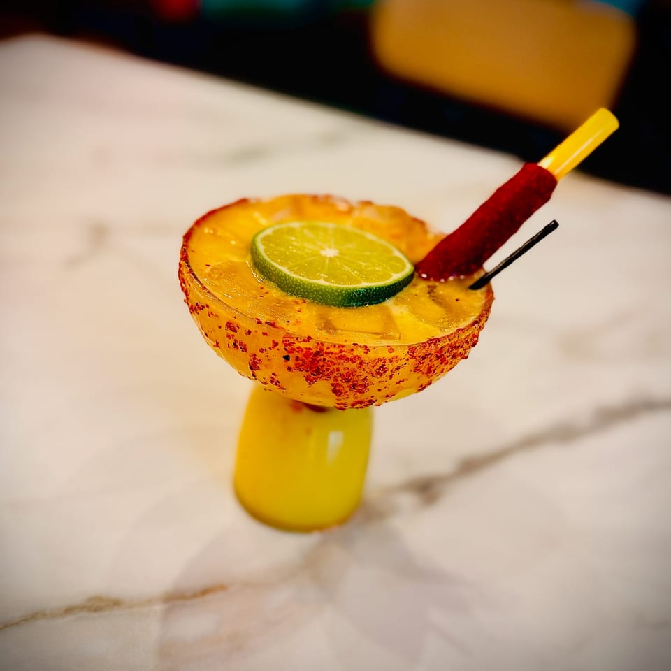 A vibrant yellow cocktail in a unique glass, garnished with a lime slice and chili-dusted straw.