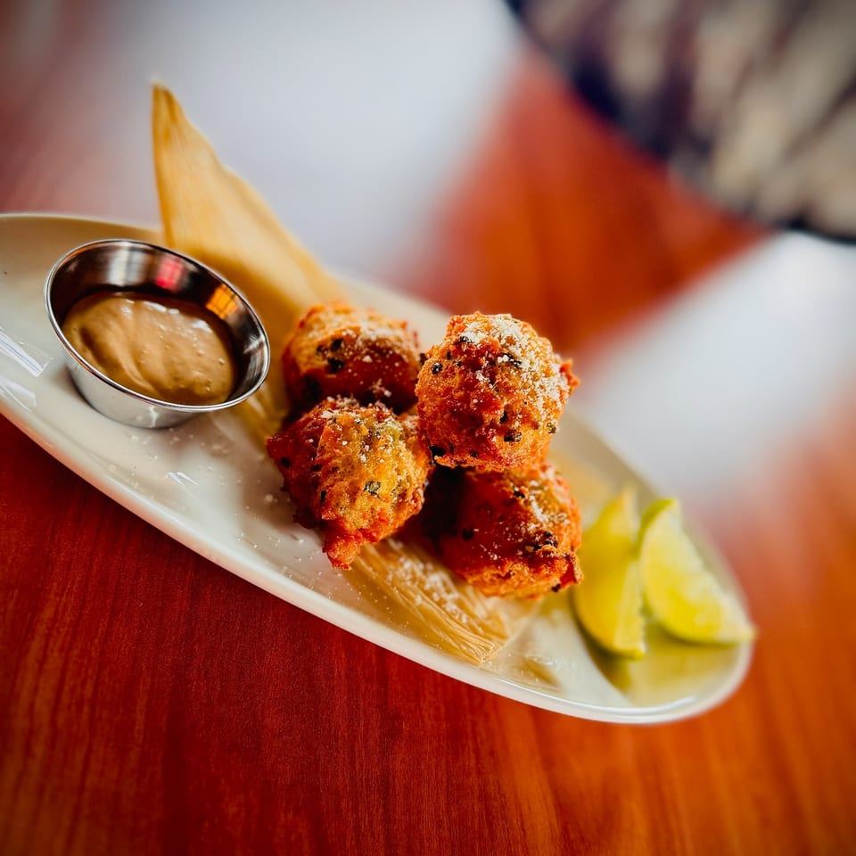 Four golden-brown hush puppies with powdered sugar, a side of sauce, and lime wedges on a white plate.