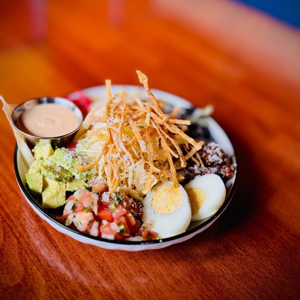 A colorful salad bowl with avocado, pico de gallo, hard-boiled eggs, crispy tortilla strips, and sauce.
