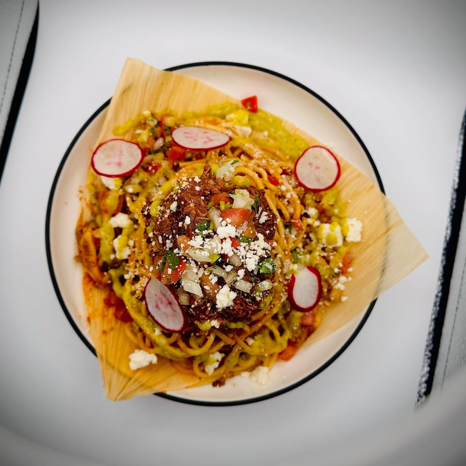 A Mexican dish with noodles, shredded meat, pico de gallo, cheese, and radishes on a corn husk.
