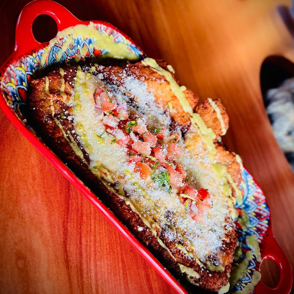 Fried plantain dish topped with white cheese, diced tomatoes, and green sauce in a red patterned bowl.