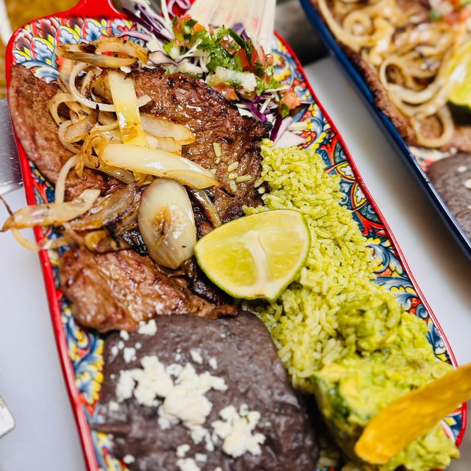Grilled steak with onions, green rice, black beans, guacamole, and lime on a colorful plate.